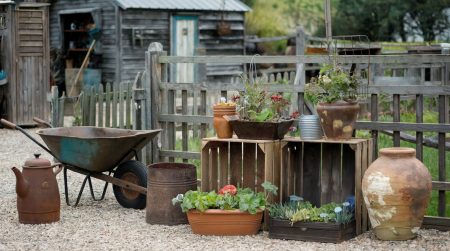 A collection of gardening containers: mason jar, rusty wheelbarrow, wooden crates and and old teapot against a backdrop of a rustic homestead.