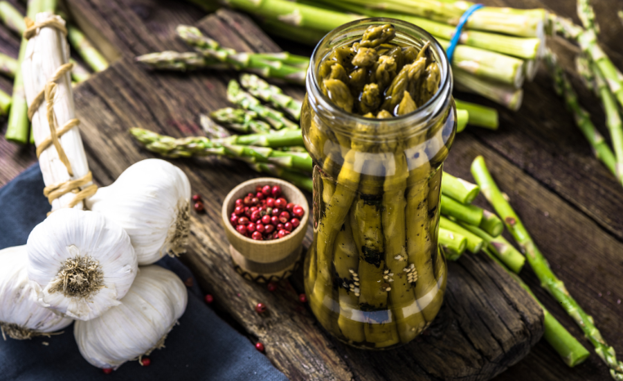 Pickled baby asparagus spears, shown stored upright on a wood board with fresh asparagus, garlic and red peppercorns.