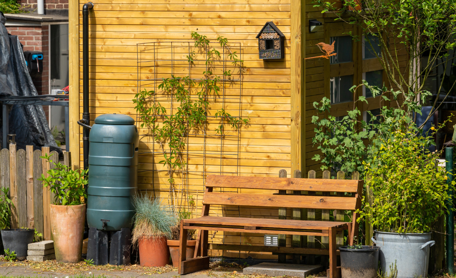 A photo of a small townhouse with a rain barrel system. The house is surrounded by a garden. There is a rain barrel connected to the downspout of the house. 