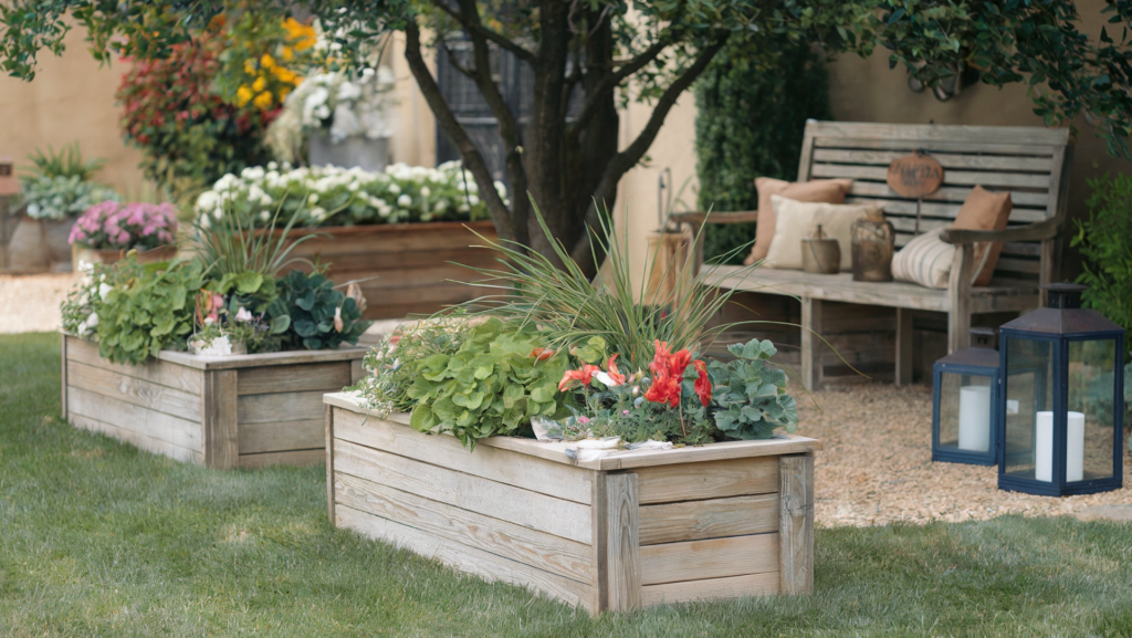 A photo of an outdoor space with barnwood planter boxes and a barnwood bench. The planter boxes contain vibrant blooms and leafy greens. The bench is placed under a tree and is surrounded by soft grass. There are rustic accessories like lanterns and pillows near the bench. 