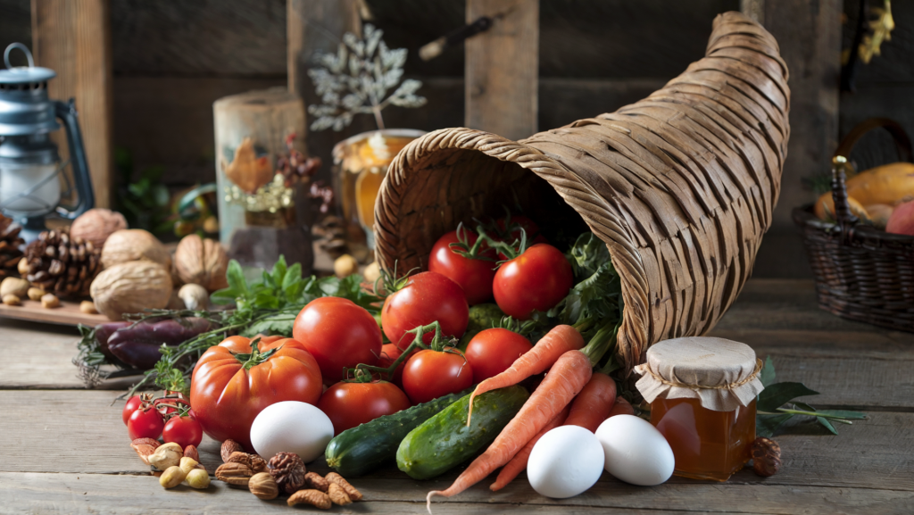 A photo of a cornucopia filled with organic tomatoes, carrots, cucumbers, fresh herbs, free-range duck eggs, and a jar of honey. The cornucopia is placed on a wooden surface. There are dried fruit, nuts, and a few seasonal decorations beside the cornucopia. The background is rustic, with wooden beams and a few items such as a lantern and a basket.