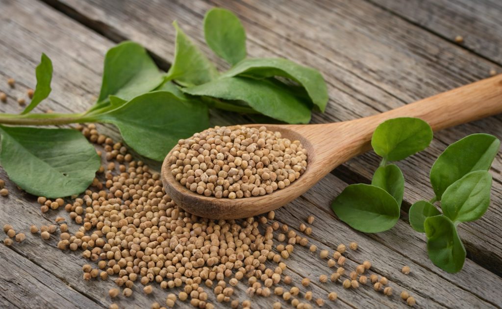 A photo of a rustic wooden table with a wooden spoon placed on it. Scattered around the spoon are numerous small, yellow fenugreek seeds. Adjacent to the seeds, there are vibrant green leaves.