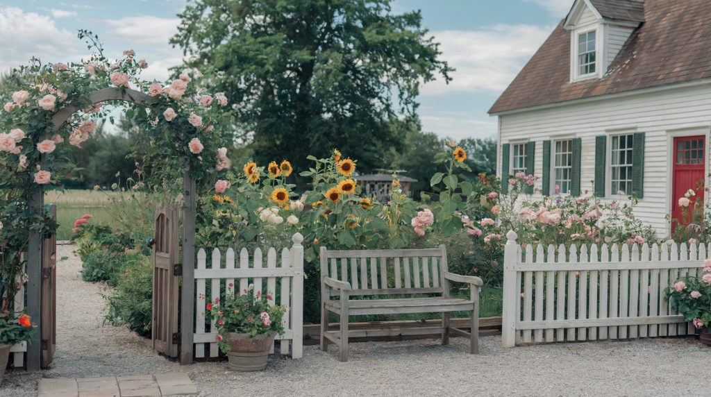 A photo of a sunny summer day on a rustic but elegant homestead with gardens. There is a white picket fence surrounding a vibrant garden filled with roses, sunflowers, and other flowers. A rustic wooden bench sits inside the fence. To the left of the garden, there is a wooden archway with pink roses. In the background, there is a white house with green shutters and a red door. The ground is covered with gravel. 