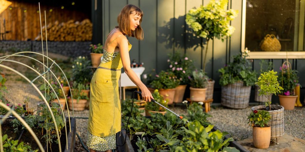 A woman in a yellow garden dress is shown watering plants in her raised bed garden.