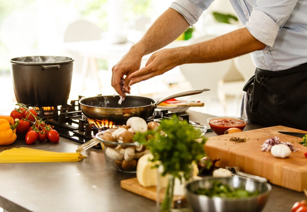 A kitchen cook is seen with a bunch of different vegetables, spices and ingredients preparing his skillet and pot for a creative one-pot meal from the pantry.