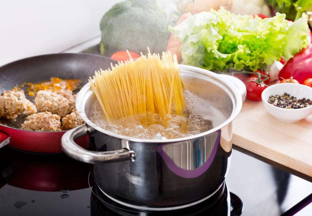 Cooking spaghetti in a pot with vegetables in the background and a skillet to one side.