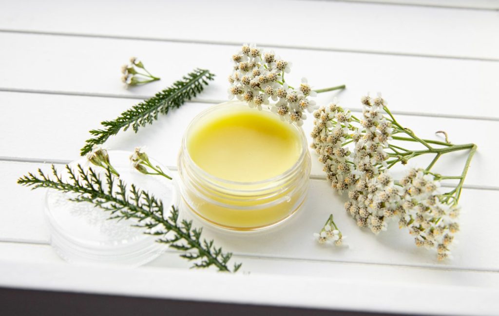 A yellow healing balm in a small glass jar is seen with sprigs of dried herbs surrounding it on a rustic white country table.